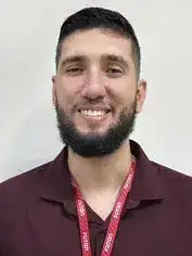 Pessoa com cabelos curtos e escuros e barba cheia bem aparada. Está sorrindo, transmite uma expressão acolhedora e veste camisa polo bordô, usando crachá com cordão vermelho. O fundo é claro e simples.