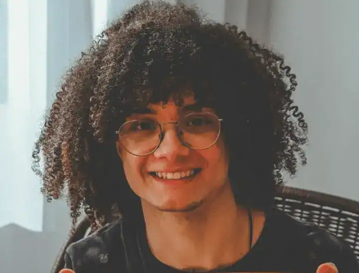 Pessoa jovem, com cabelos cacheados volumosos e escuros, usando óculos de armação redonda. Tem expressão sorridente, aparência amigável e está sentada em um ambiente interno bem iluminado, vestindo camiseta escura.