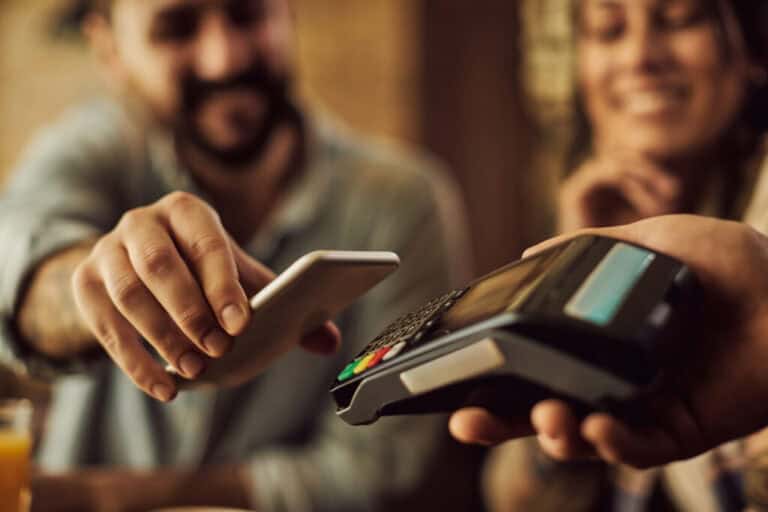 Pessoa aproxima o celular de uma maquininha para pagamento por aproximação, enquanto outra pessoa sorri ao fundo.