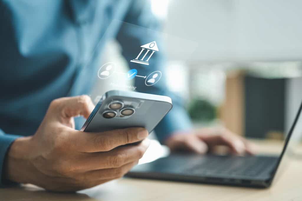 Pessoa segura um smartphone enquanto usa um notebook. Ícones de autenticação e verificação digital aparecem sobre a tela do celular.
